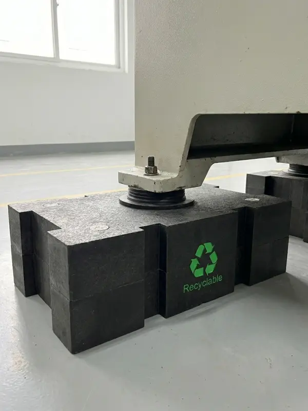 Industrial cribbing block made from fully recyclable composite material, shown holding a heavy machinery foot. The block features a prominent green recyclable symbol and text, with colorful plastic pellets and wood chips in the foreground, highlighting a sustainable alternative to traditional timber.